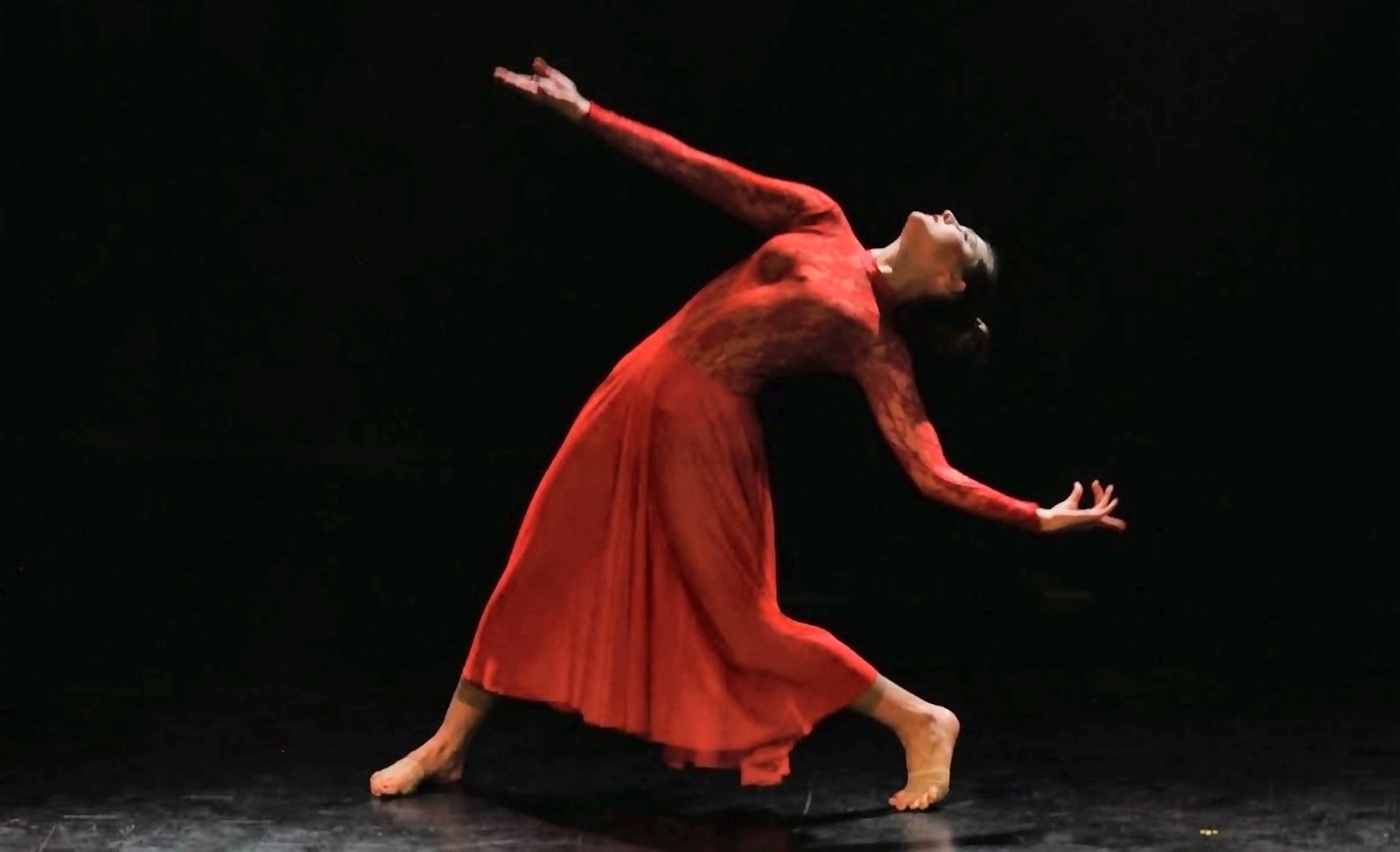 A dancer in a long red dress arching backwards on a dark stage