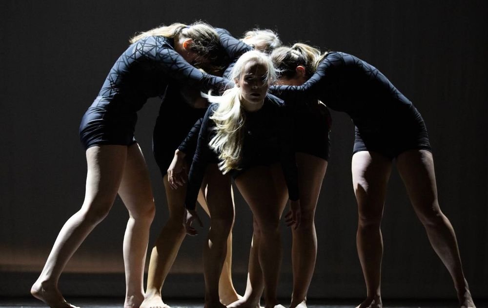 A group of dancers in black, huddled on stage